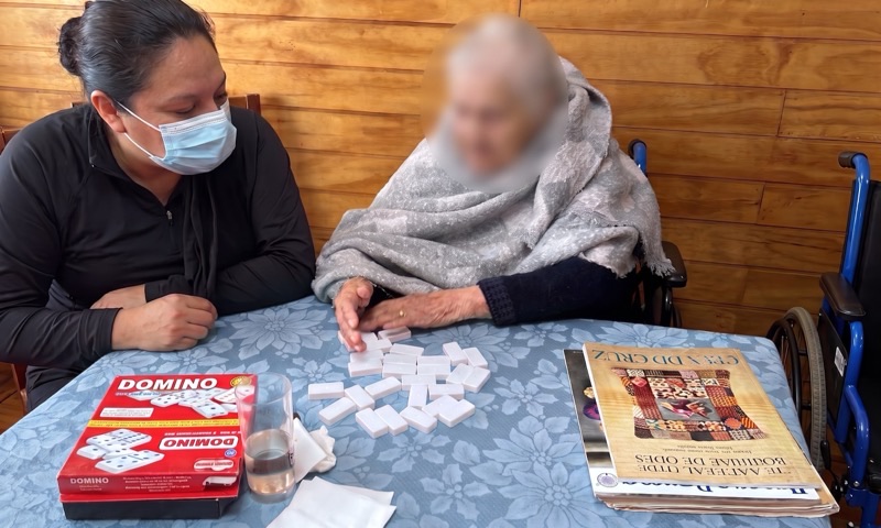 Una integrante del equipo acompaña a una residente en una partida de dominó sobre la mesa del comedor