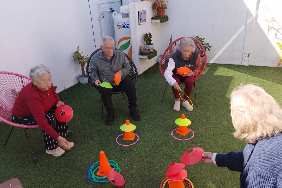 Grupo de residentes sentados en círculo en la terraza, participando en un juego de puntería con conos y aros de colores sobre el pasto