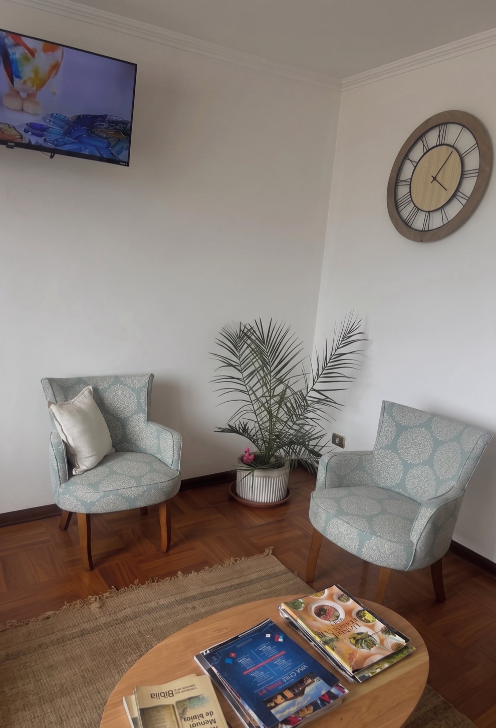 Rincón de estar de la residencia con dos sillones tapizados en verde agua, una planta y una mesa con revistas sobre una alfombra de fibra natural