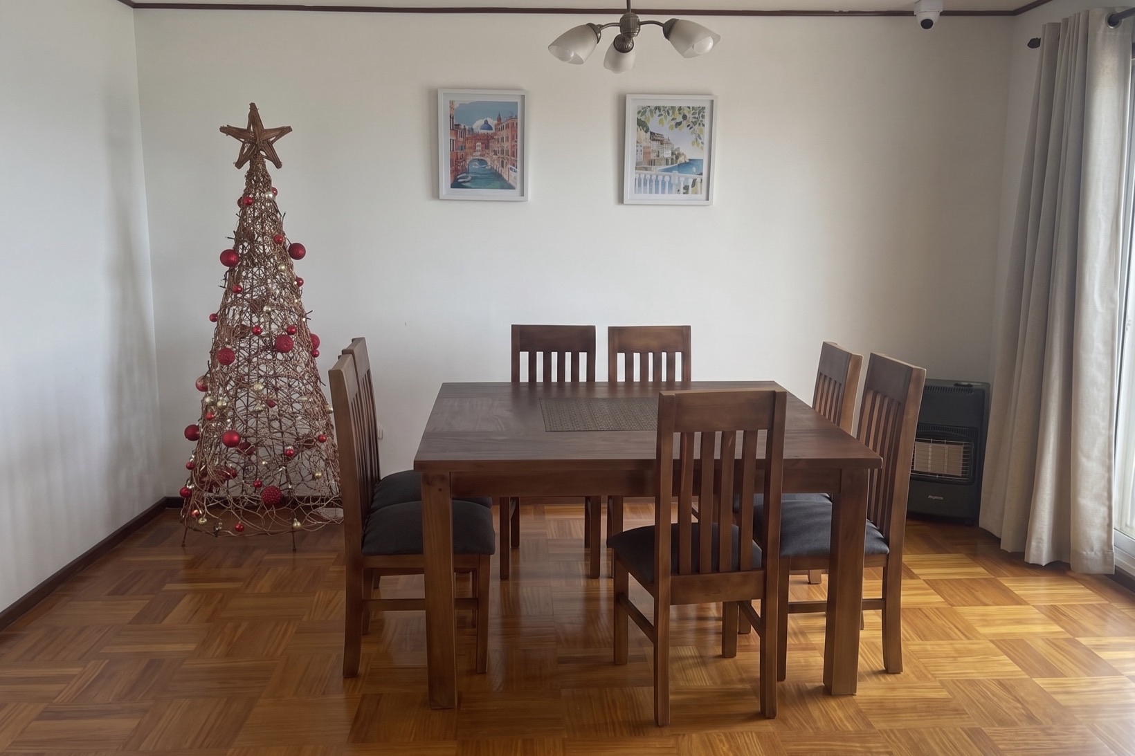 Comedor de la residencia con mesa de madera para seis personas, cuadros en la pared y cortinas que dejan entrar la luz