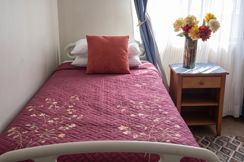 Habitación con cama individual vestida, velador de madera con flores y ventana con luz natural