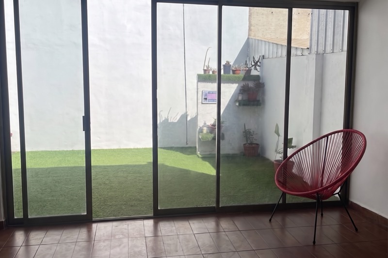 Sala con ventanal corredero que da a un patio con pasto y plantas, y un sillón junto a la ventana