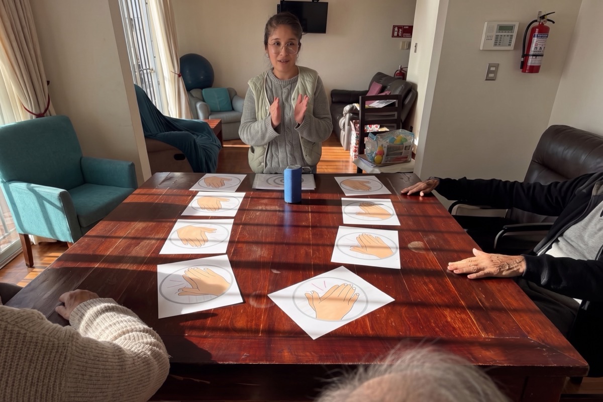 Profesional de la residencia guiando un ejercicio de coordinación con láminas de manos, mientras varios residentes participan sentados alrededor de la mesa