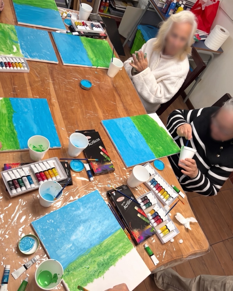 Taller de pintura: residentes trabajando en lienzos sobre una mesa con témperas, pinceles y vasos de colores